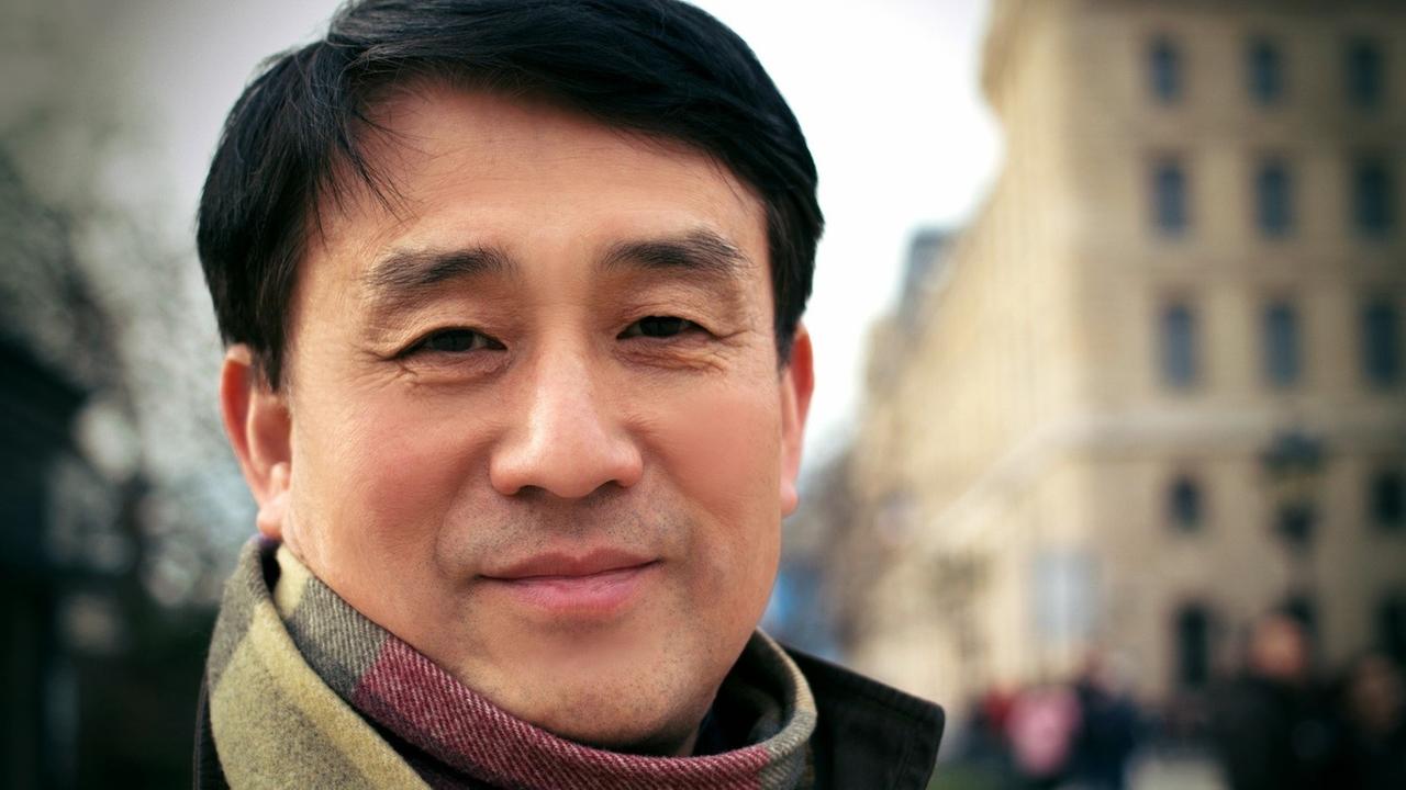 Auf dem Porträtbild ist ein lächelnder Mann mit asiatischem Aussehen vor einer Häuserzeile zu sehen.