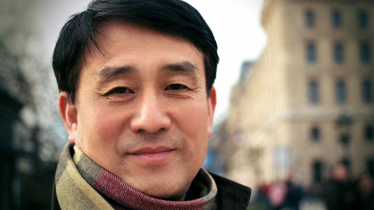 Auf dem Porträtbild ist ein lächelnder Mann mit asiatischem Aussehen vor einer Häuserzeile zu sehen. Auf dem Porträtbild ist ein lächelnder Mann mit asiatischem Aussehen vor einer Häuserzeile zu sehen.