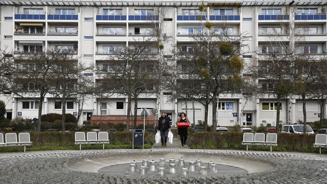 Pastor Bernd Siggelow und Sozialarbeiterin Sina Wollmann sind sehr klein vor einer Plattenbauarchitektur zu sehen. Sie tragen Tüten und laufen auf einen Platz zu. Pastor Bernd Siggelow und Sozialarbeiterin Sina Wollmann sind sehr klein vor einer Plattenbauarchitektur zu sehen. Sie tragen Tüten und laufen auf einen Platz zu.