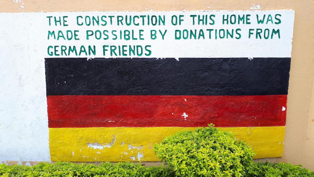 An eine Wand ist eine Deutschlandfahne gemalt, darüber die Aufschrift "The construction of this home was made possible by donations from German friends". An eine Wand ist eine Deutschlandfahne gemalt, darüber die Aufschrift "The construction of this home was made possible by donations from German friends".