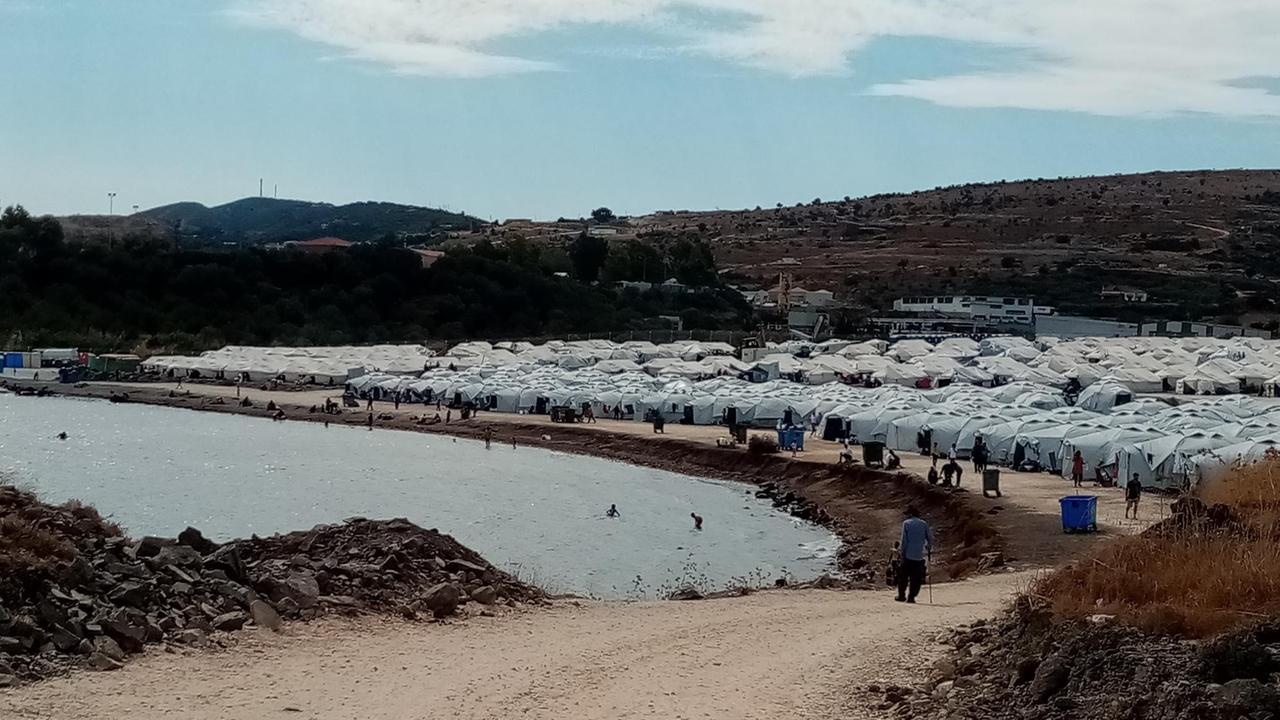 Dutzende weiße Zelte stehen direkt am Meer, in einer runden flachen Bucht. Menschen stehen im Wasser. Dutzende weiße Zelte stehen direkt am Meer, in einer runden flachen Bucht. Menschen stehen im Wasser.