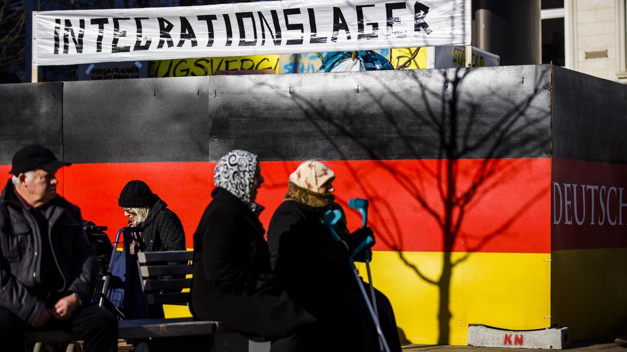 Vor einer Hauswand, in den deutschen Nationalfarben Schwarz rot gold gestrichen, sitzen zwei Frauen mit Kopftuch. Auf einem Schild ist das Wort Integrationslager zu lesen. 