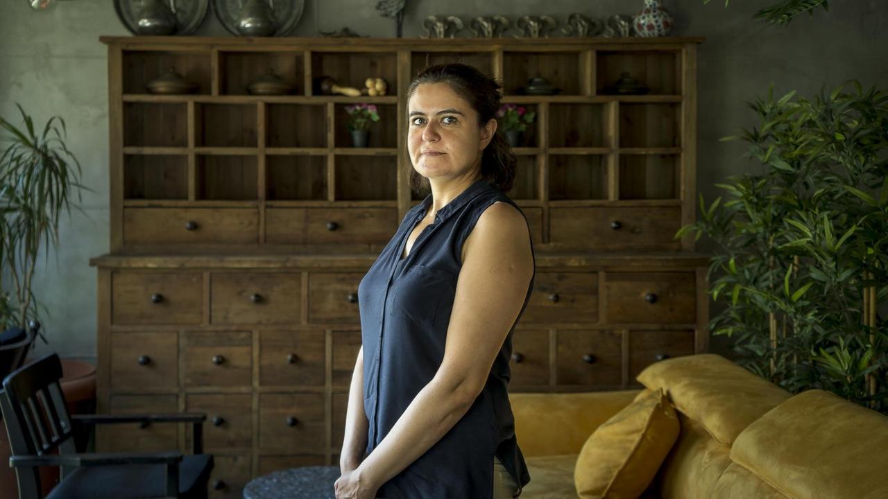 Eine Frau mit langen dunklen Haaren und blauem kurzärmligen Oberteil steht in einem dunklen Raum mit Anrichte und Sofa. Eine Frau mit langen dunklen Haaren und blauem kurzärmligen Oberteil steht in einem dunklen Raum mit Anrichte und Sofa.