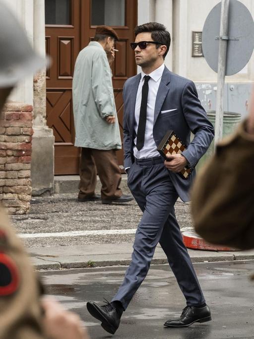 Szenenfoto aus der Spionage-Serie "Spy City", auf dem der Schauspieler Dominic Cooper in der Rolle des Agenten Fielding Scott mit einem gefalteten Schachbrett unter dem Arm an zwei Soldaten vorbei die Straße hinunter läuft.