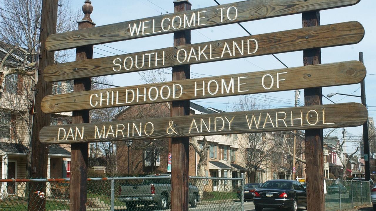 Auf den hölzernen Planken einer Gedenktafel steht die Aufschrift "Welcome to South Oakland - Childhood Home of Dan Marino & Andy Warhol" - im Hintergund sind Wohnhäuser und Teile der Straße zu sehen.