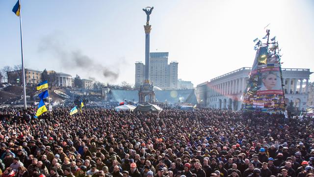 Eine große Menschenmenge auf dem Maidan-Platz in Kiew, im Hintergrund eine ukrainische Flagge an einem Mast, das Monument in der Mitte des Platzes und das von den Demonstranten errichtete Mahnmal mit dem Bild der inhaftierten Ex-Regierungschefin Julia Timoschenko