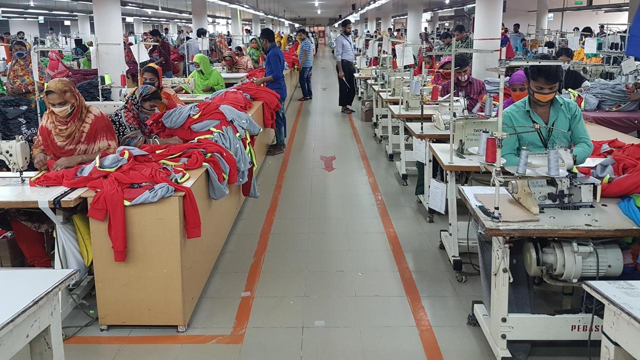 Ein Blick in eine Textilfabrik in Bangladesch. Ein Gang befindet sich zwischen einer Reihe von Tischen mit Nähmaschinen an denen Frauen sitzen und Kleidung herstellen. 