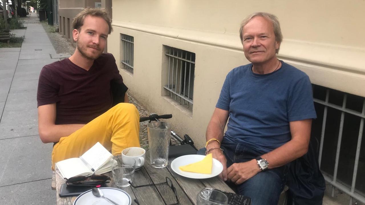 Der Künstler Kilian Jörg und der Kameramann Jan Kerhart sitzen im Außenbereich auf der Straße eines Berliner Cafés.