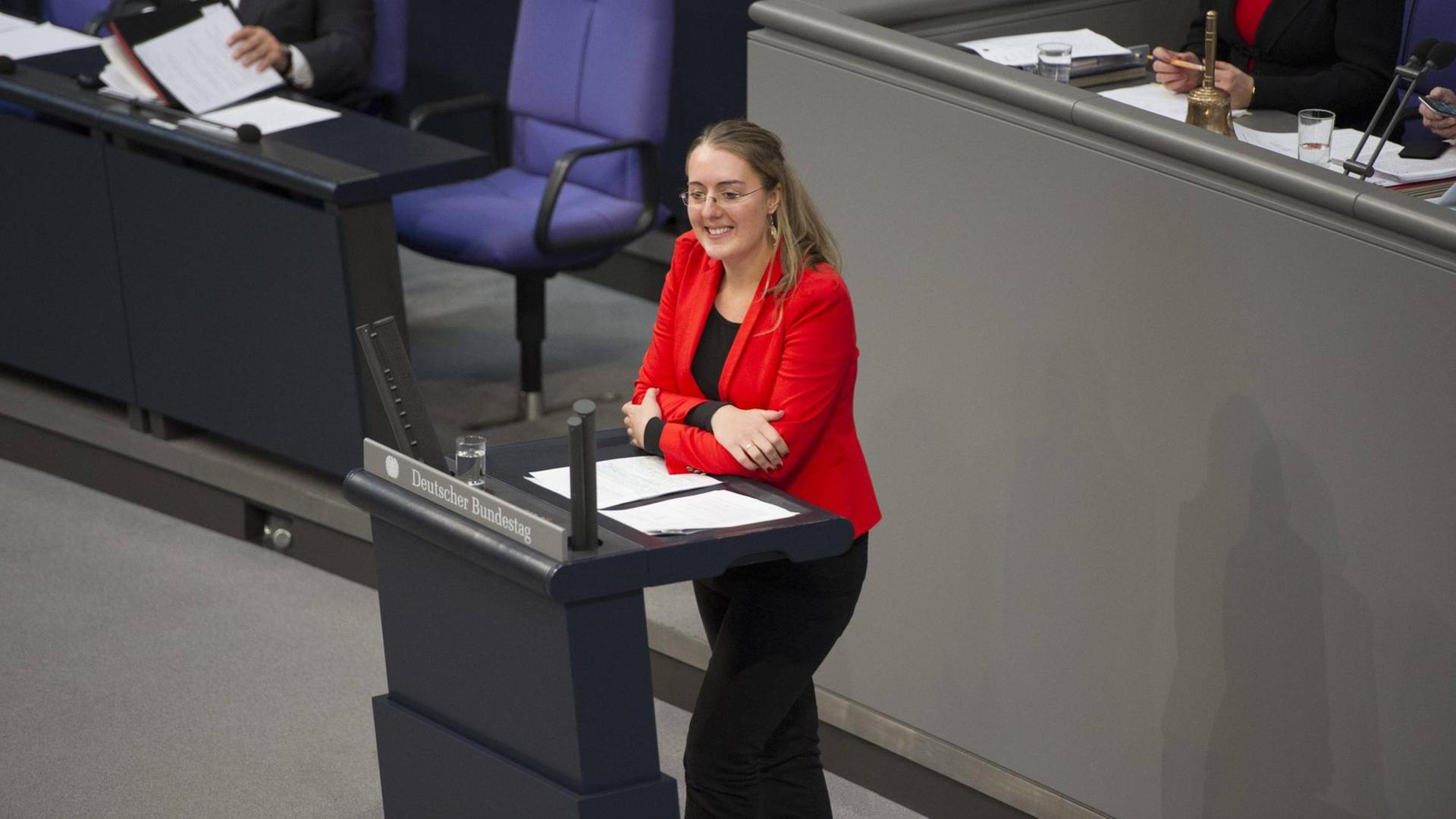 Eine Frau mit rotem Jacket hält kurz inne, während einer Rede am Pult stehend, im Deutschen Bundestag