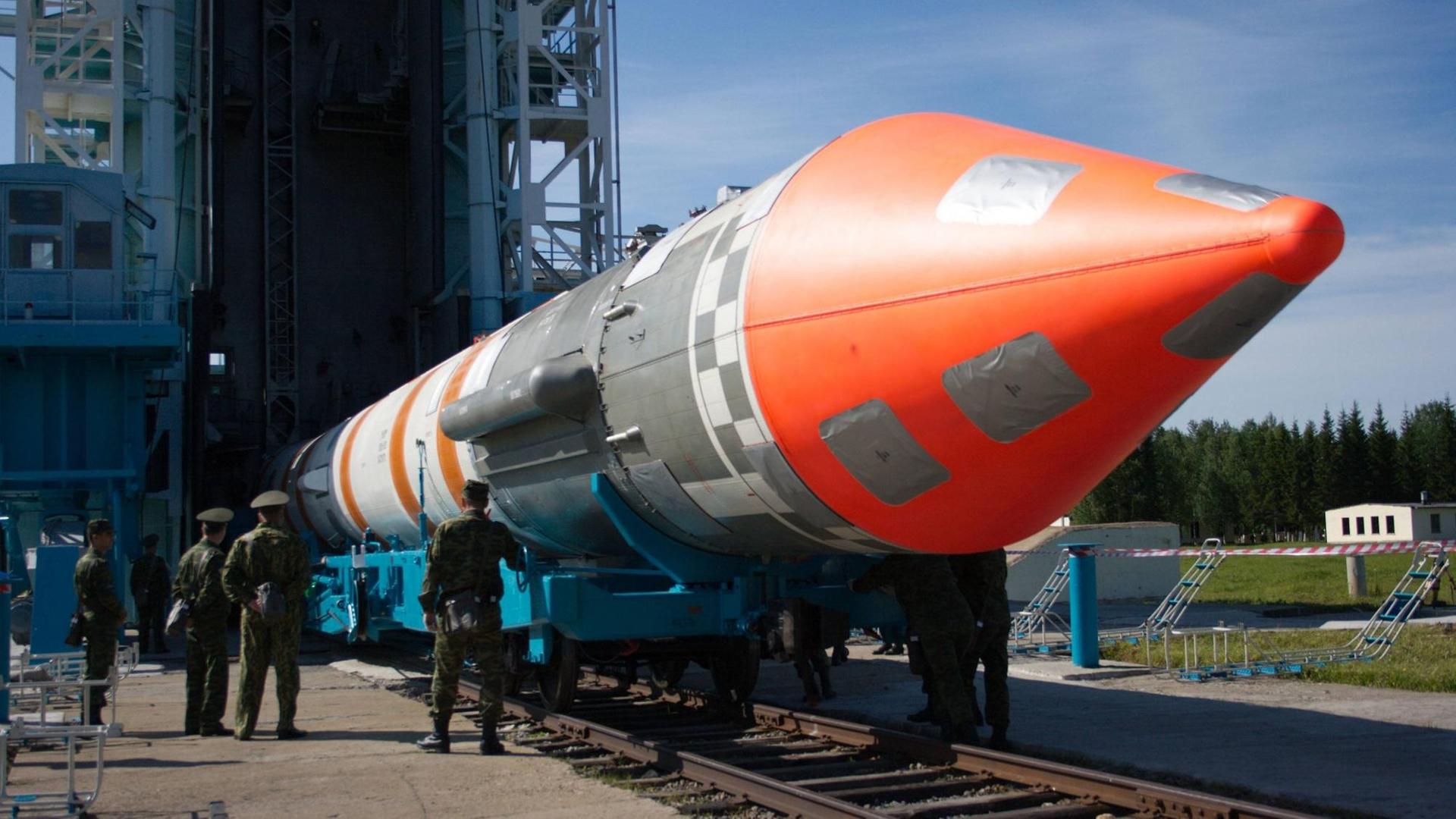 Eine riesige, rot-silberne Cosmos-3 Rakete liegt auf dem Boden vor einer Abschussrampe. Davor stehen drei russische Soldaten in Uniform. Eine riesige, rot-silberne Cosmos-3 Rakete liegt auf dem Boden vor einer Abschussrampe. Davor stehen drei russische Soldaten in Uniform.