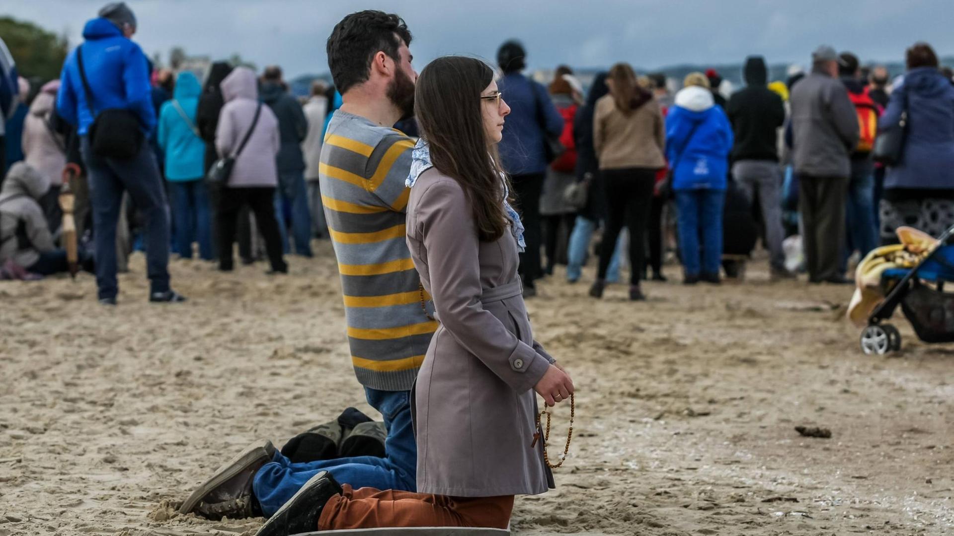 Zwei Gläubige knieen am Strand, dahinter sind weitere Gläubige am Küstenabschnitt versammelt Zwei Gläubige knieen am Strand, dahinter sind weitere Gläubige am Küstenabschnitt versammelt
