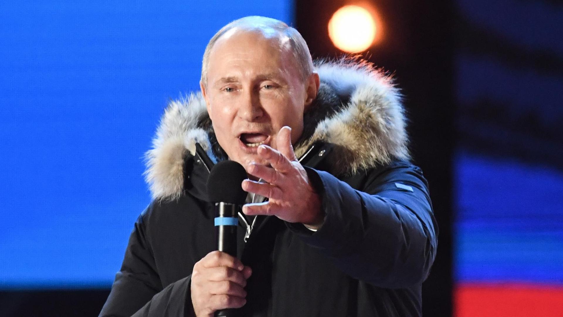 Putin steht in Winterjacke auf einer Bühne im abendlichen Moskau Putin steht in Winterjacke auf einer Bühne im abendlichen Moskau