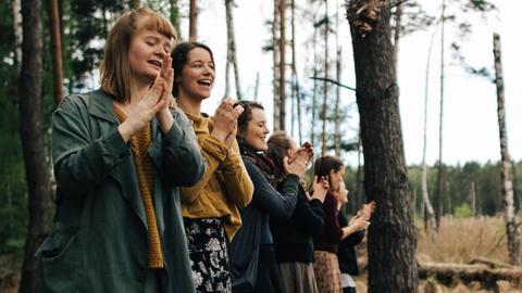 Sechs junge Frauen stehen in einem Wald und singen.