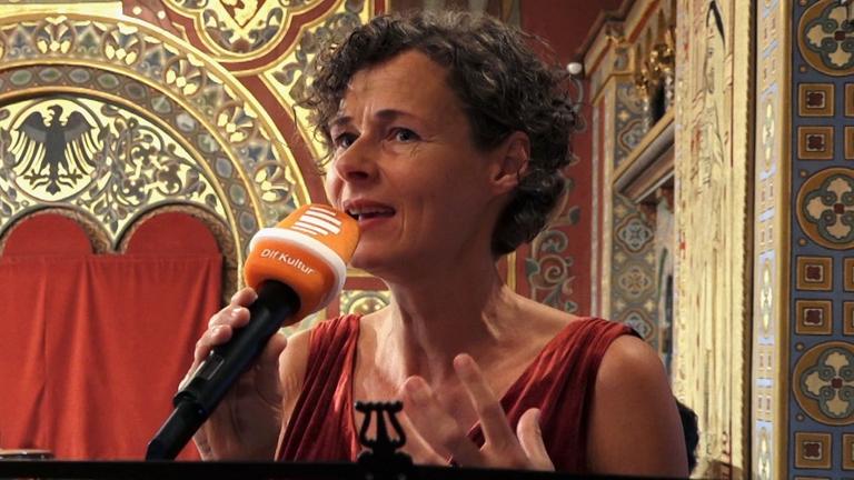 Die Moderatorin spricht  im Palas in ihr Mikrofon mit dem Logo vom Deutschlandfunk Kultur.