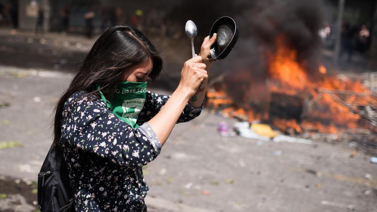 Gewaltsame Proteste in Santiago de Chile am 23. Oktober 2019. Eine Frau mit einem Tuch vor dem Gesicht schlägt mit einem Löffel auf einen Topf. Im Hintergrund brennen Barrikaden. Gewaltsame Proteste in Santiago de Chile am 23. Oktober 2019. Eine Frau mit einem Tuch vor dem Gesicht schlägt mit einem Löffel auf einen Topf. Im Hintergrund brennen Barrikaden.