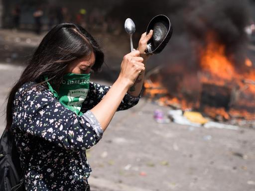 Gewaltsame Proteste in Santiago de Chile am 23. Oktober 2019. Eine Frau mit einem Tuch vor dem Gesicht schlägt mit einem Löffel auf einen Topf. Im Hintergrund brennen Barrikaden.
