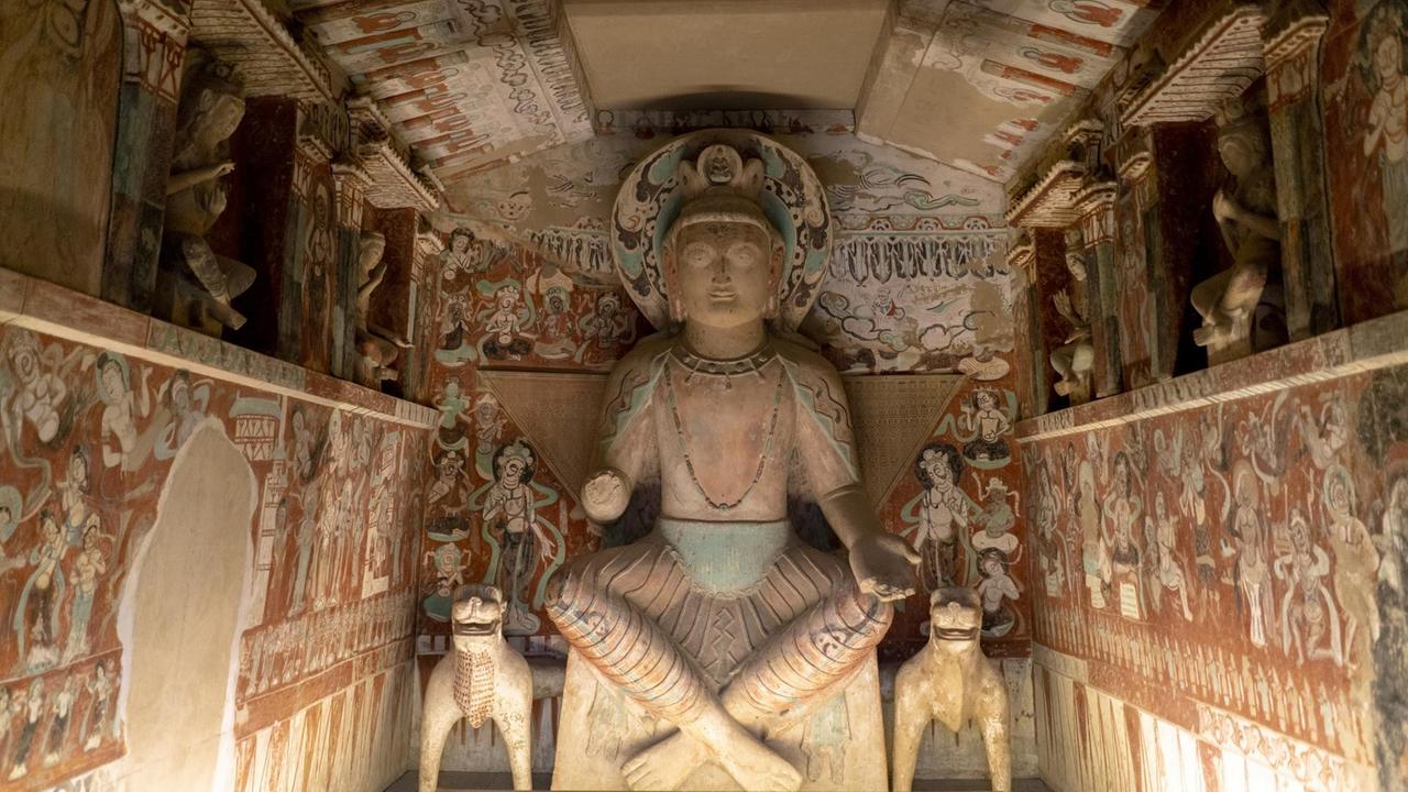Buddhastatue in einer Höhle, umgeben von farbenfrohen Wandgemälden.
