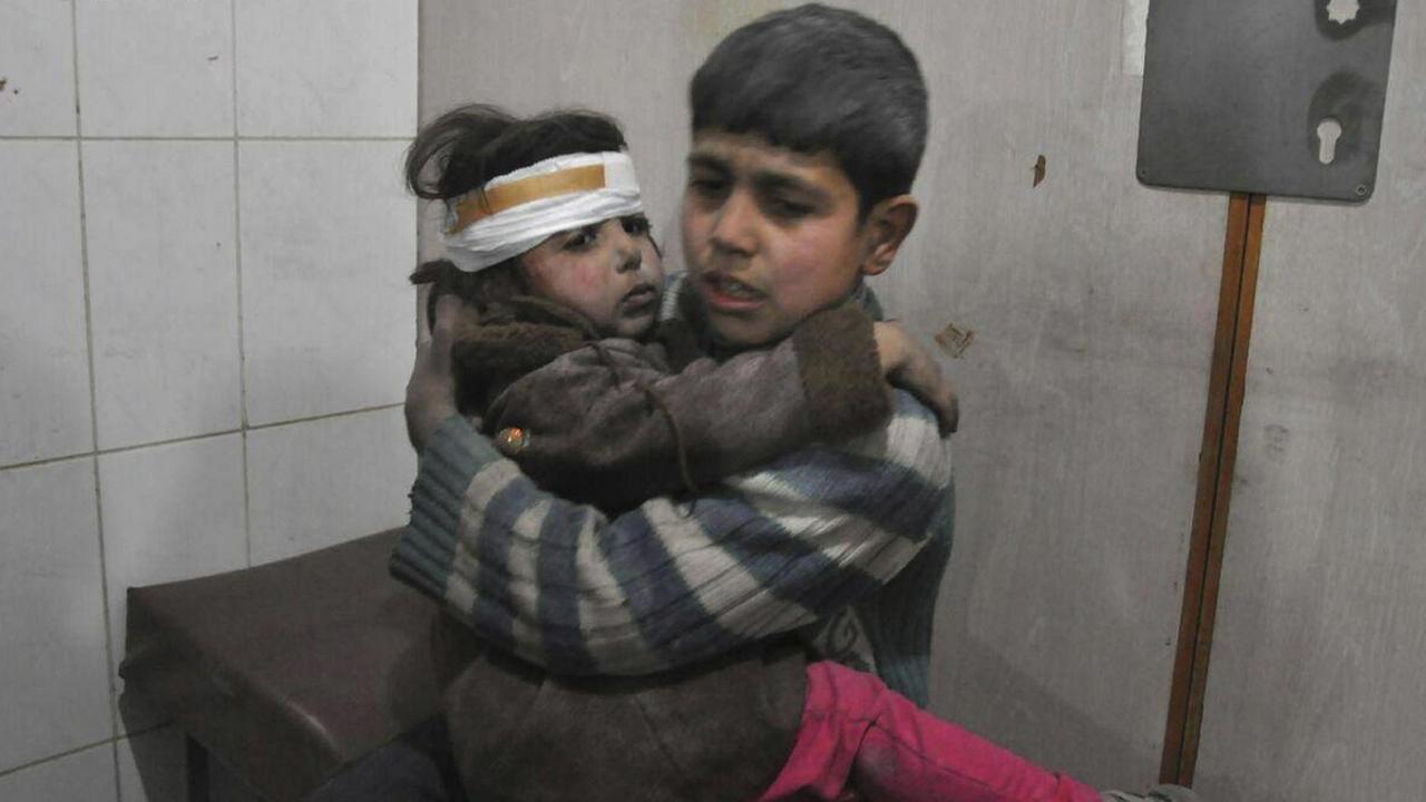 Der etwa zehnjährige Junge sitzt in einem Raum auf einer Bahre und hält sein kleines Geschwisterkind im Arm, dessen Kopf verbunden ist. 