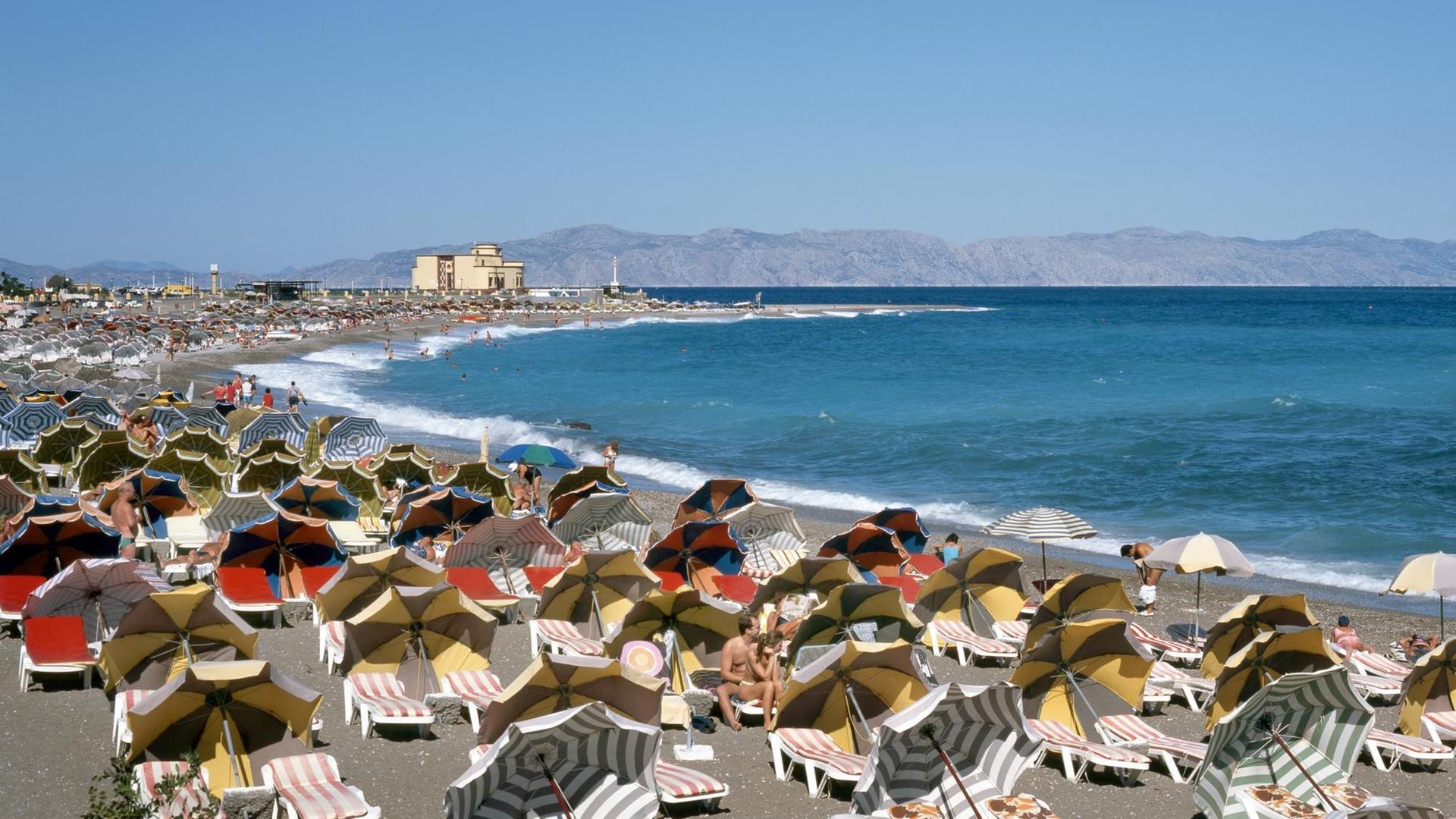 Touristen sitzen unter Sonnenschirmen und auf Liegen am Strand von Rhodos-Stadt.