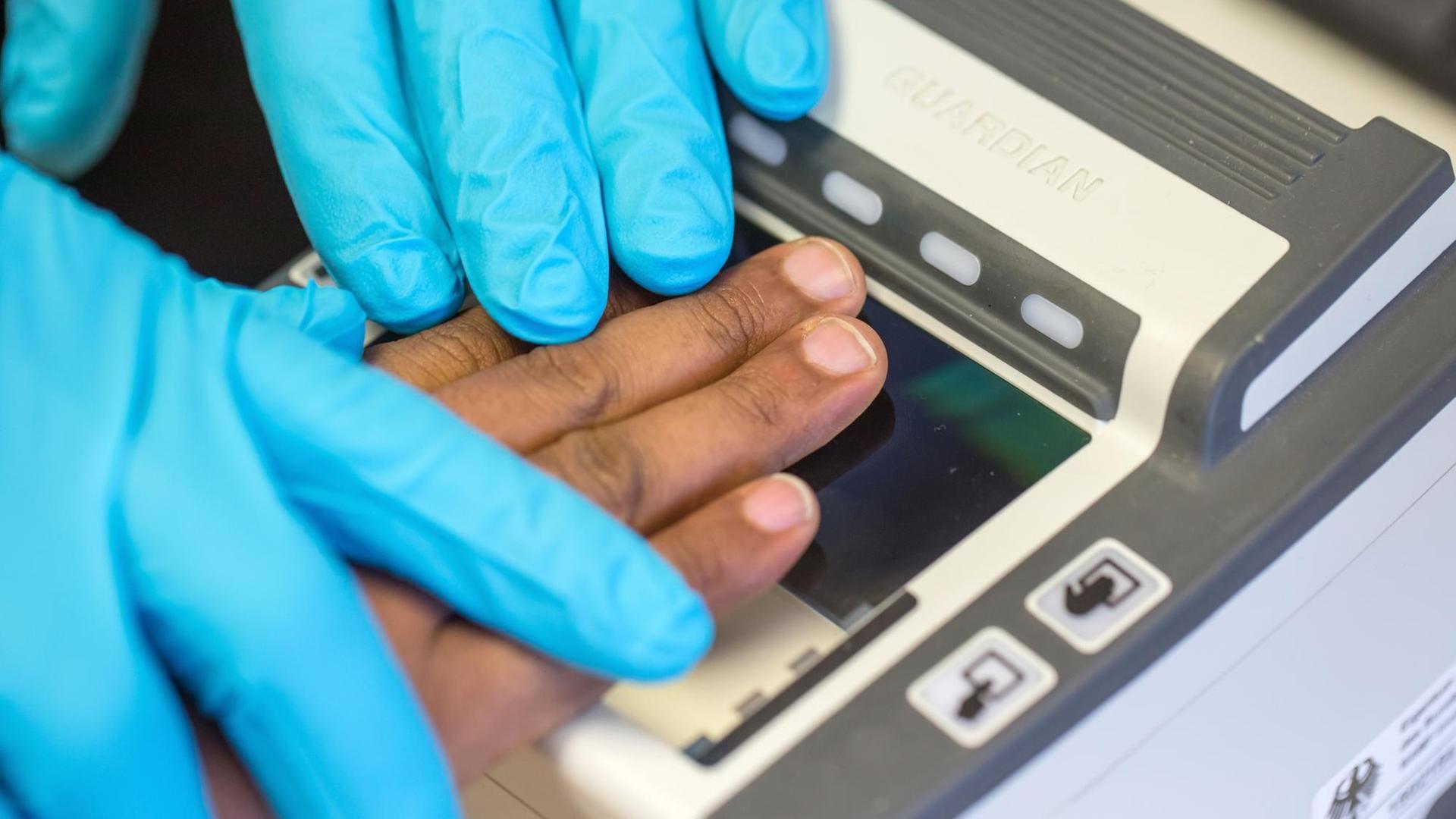 Zwei Hände in Gummihandschuhen halten die Hand eines schwarzen Flüchtlings an einGgerät zur Registrierung der Fingerabdrücke. Zwei Hände in Gummihandschuhen halten die Hand eines schwarzen Flüchtlings an einGgerät zur Registrierung der Fingerabdrücke.