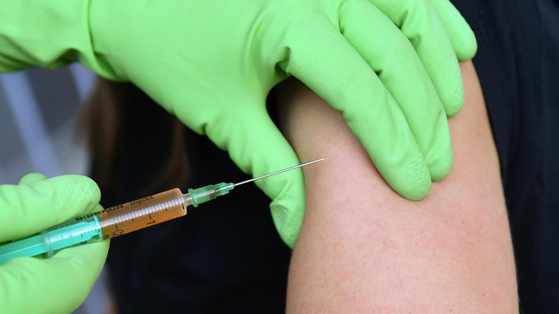 Zwei Hände in Schutzhandschutzen drücken einen Oberarm zusammen und setzen eine Spritze an. Zwei Hände in Schutzhandschutzen drücken einen Oberarm zusammen und setzen eine Spritze an.