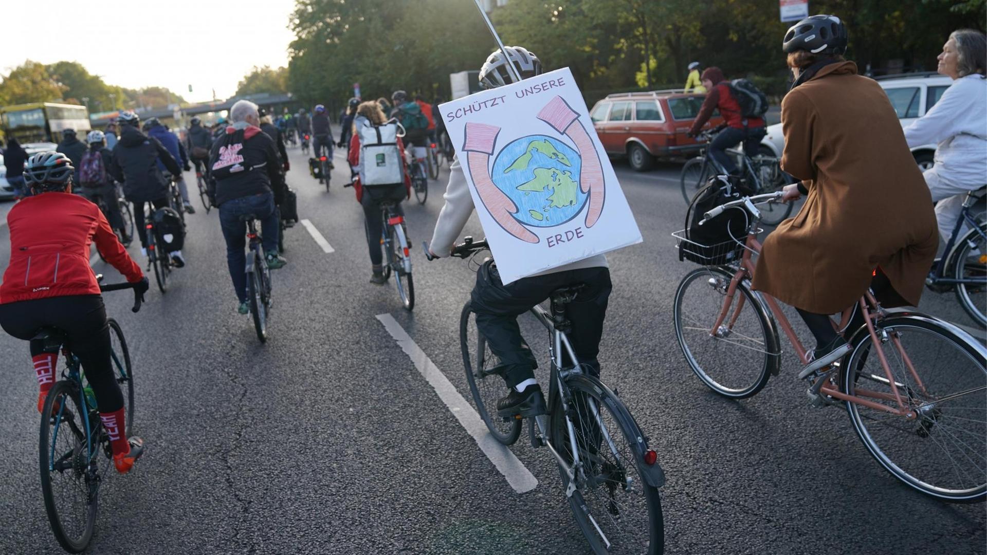 Mehrere Fahrradfahrer auf einer zweispurigen Straße im Sonnenaufgang. Einer trägt auf dem Rücken ein Plakat mit der Aufschrift "Schützt unsere Erde". Mehrere Fahrradfahrer auf einer zweispurigen Straße im Sonnenaufgang. Einer trägt auf dem Rücken ein Plakat mit der Aufschrift "Schützt unsere Erde".