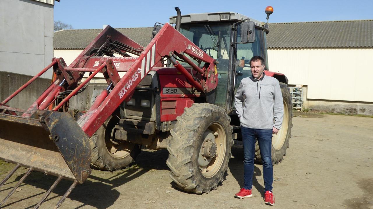 Ein junger Mann in Jeans und roten Schuhen steht auf einem Hof neben einem roten Traktor mit ausgefahrener Schaufel. Ein junger Mann in Jeans und roten Schuhen steht auf einem Hof neben einem roten Traktor mit ausgefahrener Schaufel.