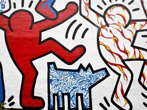 Das Bild zeigt einen Ausschnitt von Harings berühmten Strichmännchen als Wandmalerei. Links ein rotes und rechts ein weißes Strichmännchen mit rot-gelben Streifen. In der Mitte unten ist ein kleiner Hund mit blau-weißem Muster zu sehen.