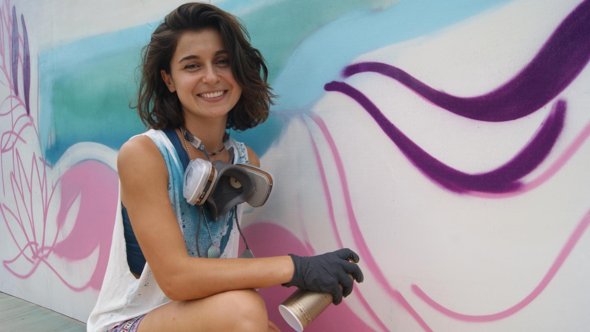 Auf dem Bild ist die Künstlerin Gamze Yalçın zu sehen. Sie lächelt in die Kamera und hält eine Sprühdose in der Hand. Rechts von ihr ist ein Graffiti zu sehen, an dem sie gerade arbeitet. Auf dem Bild ist die Künstlerin Gamze Yalçın zu sehen. Sie lächelt in die Kamera und hält eine Sprühdose in der Hand. Rechts von ihr ist ein Graffiti zu sehen, an dem sie gerade arbeitet.