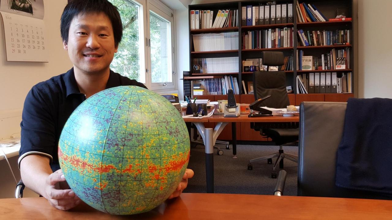 Eiichiro Komatsu sitzt an einem Schreibtisch und hält einen Globus in den Händen Eiichiro Komatsu sitzt an einem Schreibtisch und hält einen Globus in den Händen