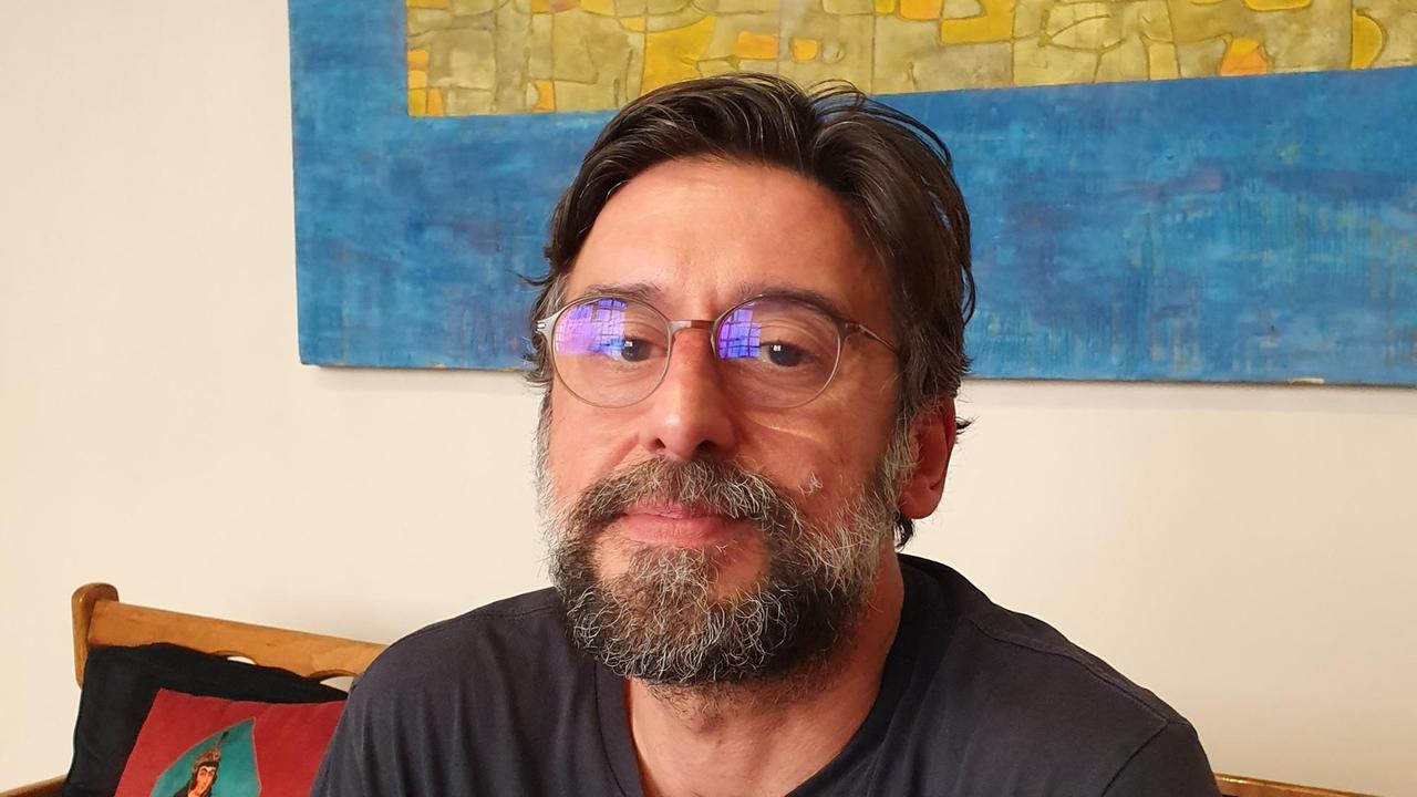 Ramin Sadighi - mit angegrautem Vollbart und Brille - in seiner Wohnung auf einem Sofa vor einem großen Bild.