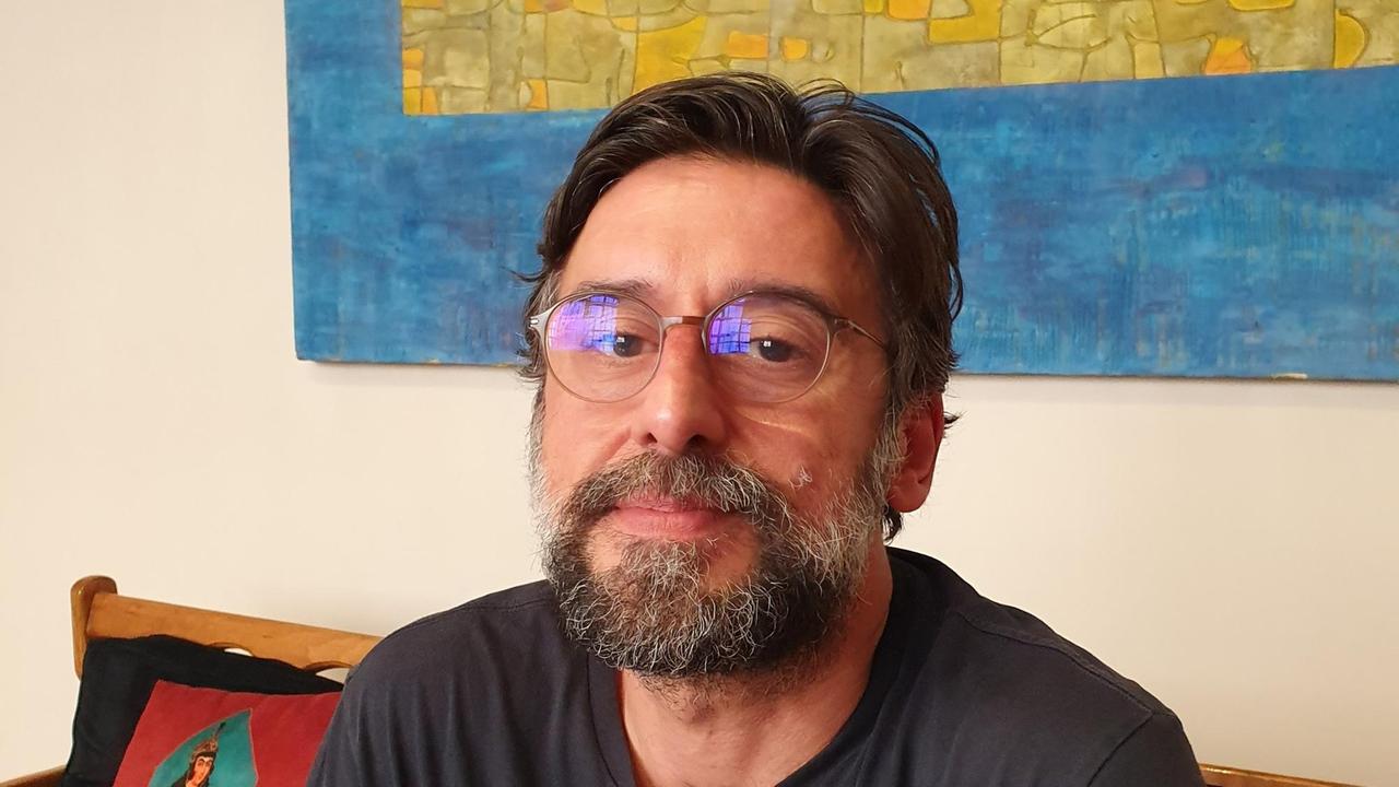 Ramin Sadighi - mit angegrautem Vollbart und Brille - in seiner Wohnung auf einem Sofa vor einem großen Bild. Ramin Sadighi - mit angegrautem Vollbart und Brille - in seiner Wohnung auf einem Sofa vor einem großen Bild.