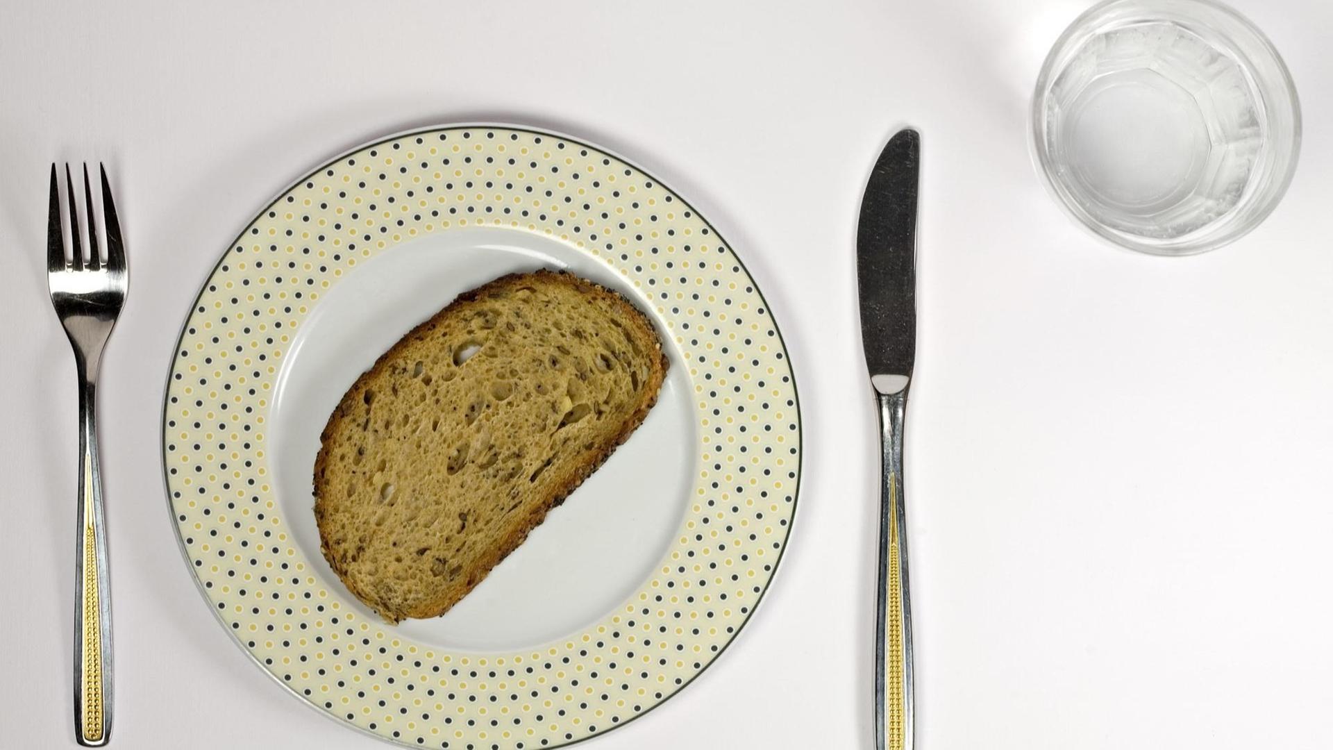 Neben einem Teller, auf dem eine Scheibe Brot liegt, liegen Messer und Gabel. Zudem steht ein mit Wasser gefülltes Glas dabeil Neben einem Teller, auf dem eine Scheibe Brot liegt, liegen Messer und Gabel. Zudem steht ein mit Wasser gefülltes Glas dabeil