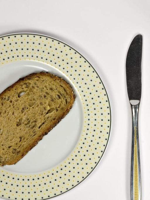 Neben einem Teller, auf dem eine Scheibe Brot liegt, liegen Messer und Gabel. Zudem steht ein mit Wasser gefülltes Glas dabeil