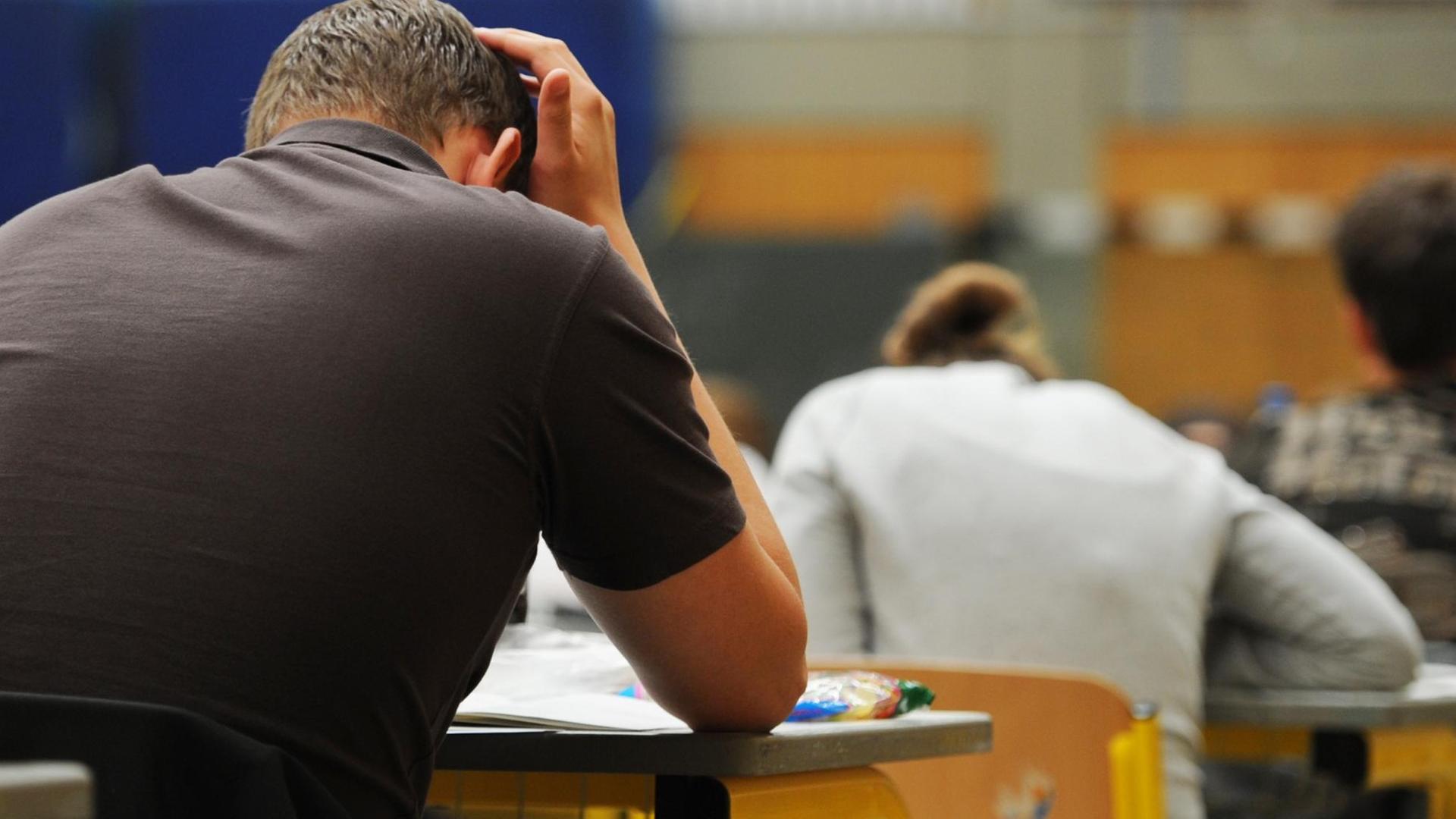 Zwei von schräg hinten aufgenommene Schüler sitzen während der Abiturprüfung an ihren Tischen in nachdenklicher Pose. Zwei von schräg hinten aufgenommene Schüler sitzen während der Abiturprüfung an ihren Tischen in nachdenklicher Pose.