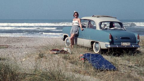 Eine Frau steht in 50er-Bademode an einem Wagen aus den 50ern. Der Wagen steht am Strand, am Meer.