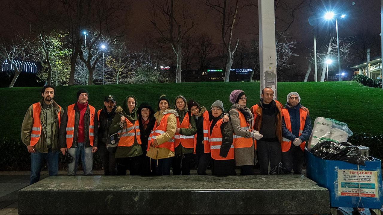 Zwölf Menschen jeden Alters und Geschlechts in orangenen Warnwesten posieren vor einer Hecke und hinter einem großen Stein bei Nacht. Zwölf Menschen jeden Alters und Geschlechts in orangenen Warnwesten posieren vor einer Hecke und hinter einem großen Stein bei Nacht.