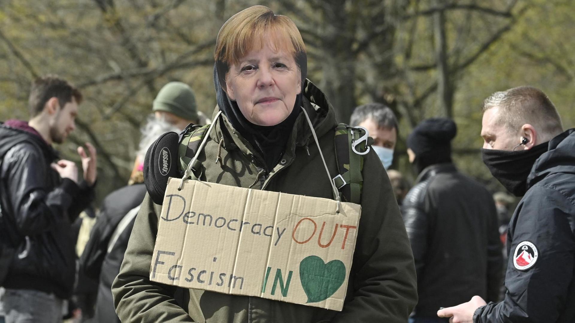 Ein Mann auf einer Querdenker Demonstration trägt eine Angela Merkel Maske und hat ein Schild um den Hals hängen: "Democracy out, Fascism in". Berlin 13. April 2021.