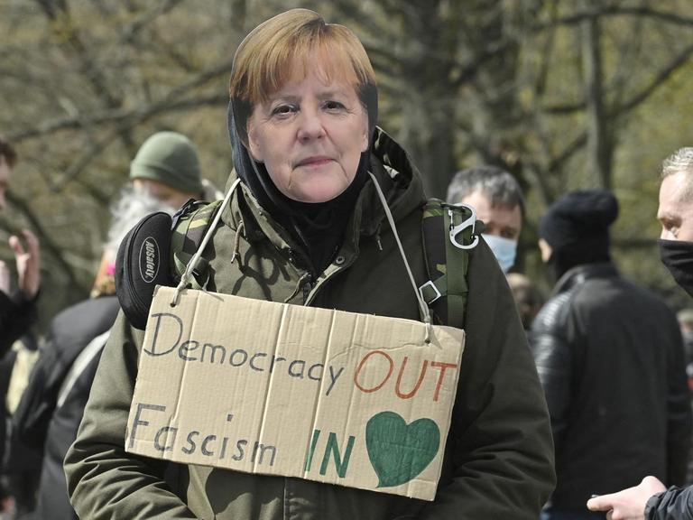 Ein Mann auf einer Querdenker Demonstration trägt eine Angela Merkel Maske und hat ein Schild um den Hals hängen: "Democracy out, Fascism in". Berlin 13. April 2021.