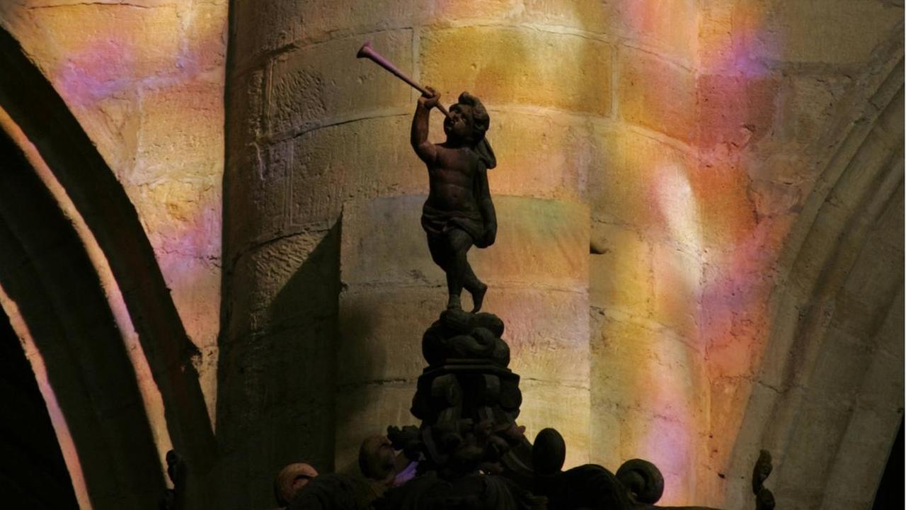 Ein Trompete spielender Steinengel steht vor einer von bunten Glasfenstern beleuchteten Wand. Ein Trompete spielender Steinengel steht vor einer von bunten Glasfenstern beleuchteten Wand.