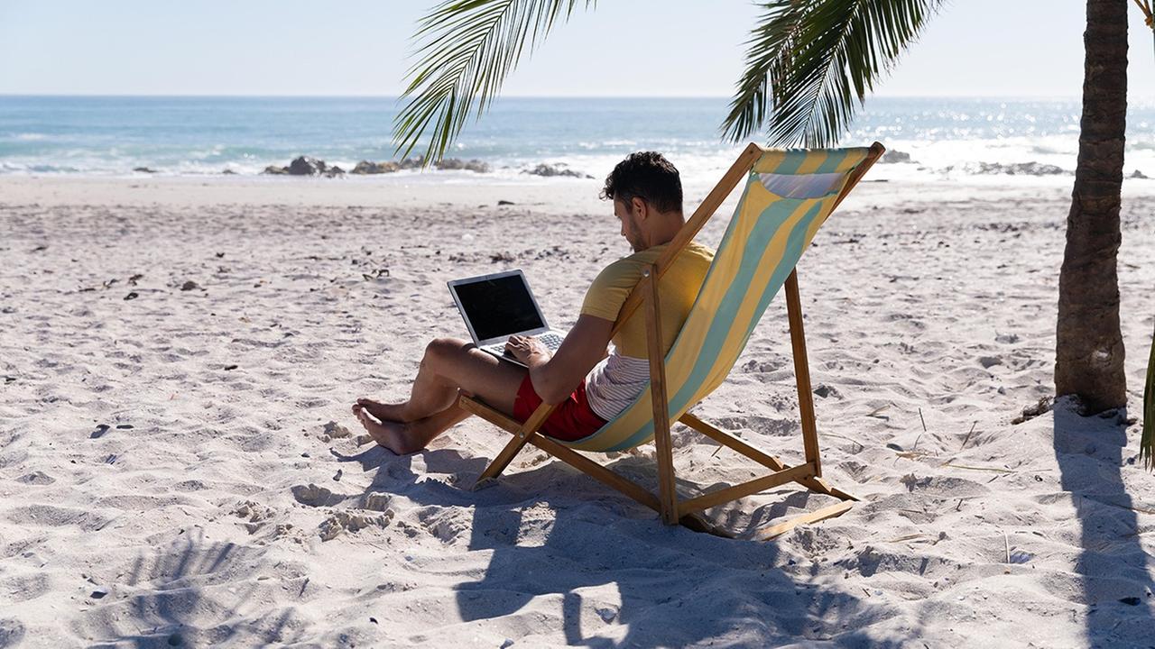 Ein Mann sitzt unter einer Palme an einem Laptop am Strand. Ein Mann sitzt unter einer Palme an einem Laptop am Strand.
