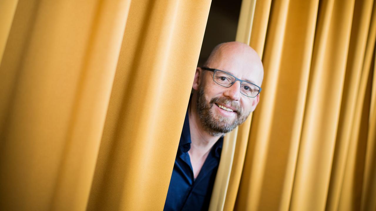 Der Intendant der Oper Wuppertal, Berthold Schneider, guckt zwischen zwei gelben langen Vorhängen in die Kamera. Der Intendant der Oper Wuppertal, Berthold Schneider, guckt zwischen zwei gelben langen Vorhängen in die Kamera.
