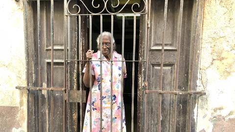 Eine ältere Kubanerin steht in der Altstadt von Havanna vor ihrer etwas baufälligen Haustür und einer Hauswand, von der der Putz bröckelt, und greift mit der rechten Hand an das hohe Gitter, das sie von der Straße trennt.