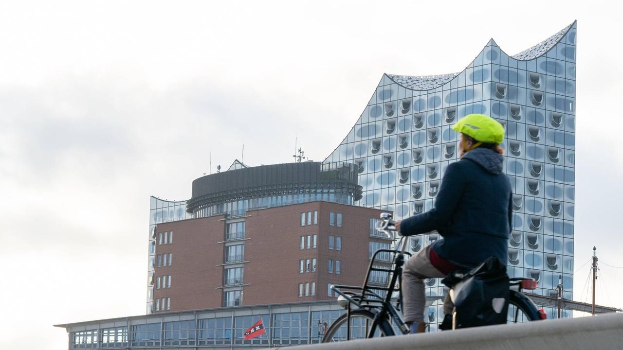 Eine fahrende Radfahrerin mit Helm ist von hinten zu sehen, im Hintergrund die Elbphilharmonie. Eine fahrende Radfahrerin mit Helm ist von hinten zu sehen, im Hintergrund die Elbphilharmonie.