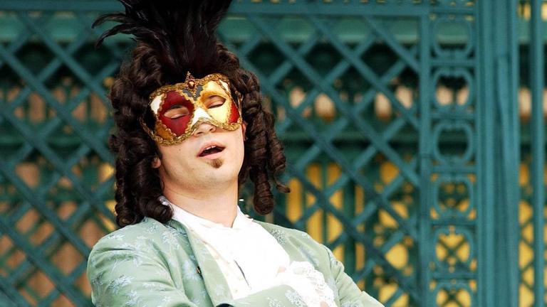 Ein Sänger mit Perücke, Maske und Rokoko-Mantel steht im Freien und singt. Ein Sänger mit Perücke, Maske und Rokoko-Mantel steht im Freien und singt.
