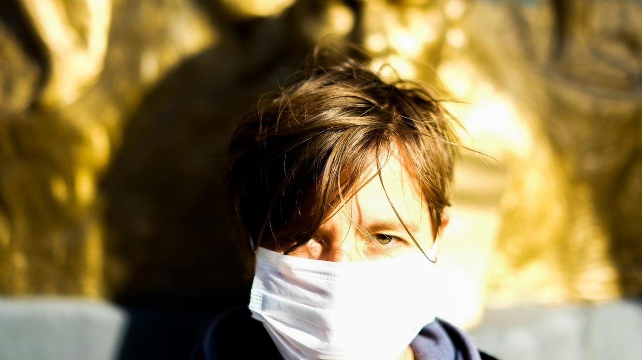 Eine dunkelblonde Frau mit weißer Maske vor Mund und Nase sitzt im Sonnenschein vor einer Skulptur und blickt in die Kamera
Eine dunkelblonde Frau mit weißer Maske vor Mund und Nase sitzt im Sonnenschein vor einer Skulptur und blickt in die Kamera
