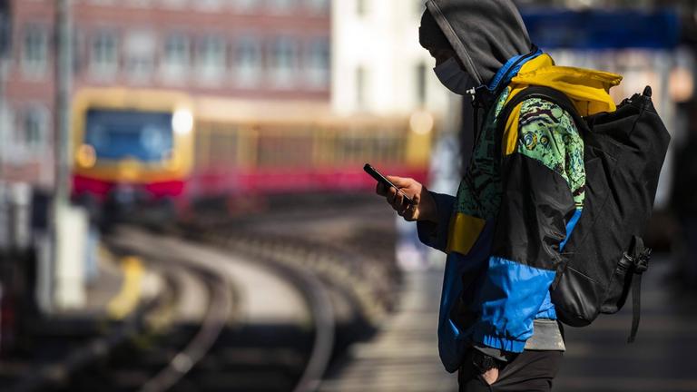 Ein Jugendlicher steht mit Mundschutzmaske auf einem S-Bahnsteig in Berlin und blickt auf sein Handy.