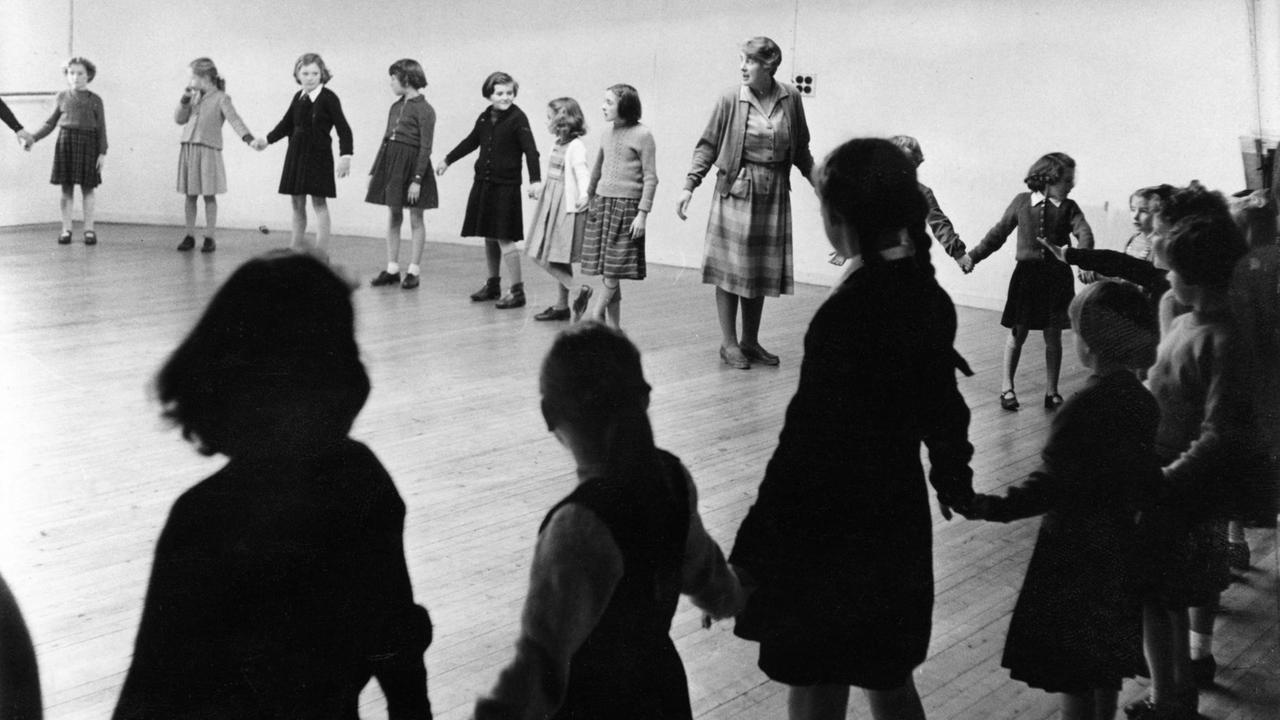 Schülerinnen und ihre Lehrerin stehen im Kreis und halten einander an den Händen. Schülerinnen und ihre Lehrerin stehen im Kreis und halten einander an den Händen.