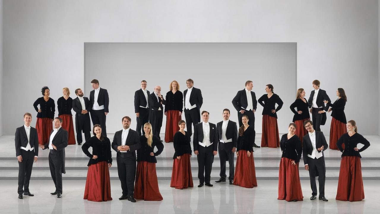 Der Chor steht in schwarz-roter Chorkleidung in einem hellen Raum. Der Chor steht in schwarz-roter Chorkleidung in einem hellen Raum.