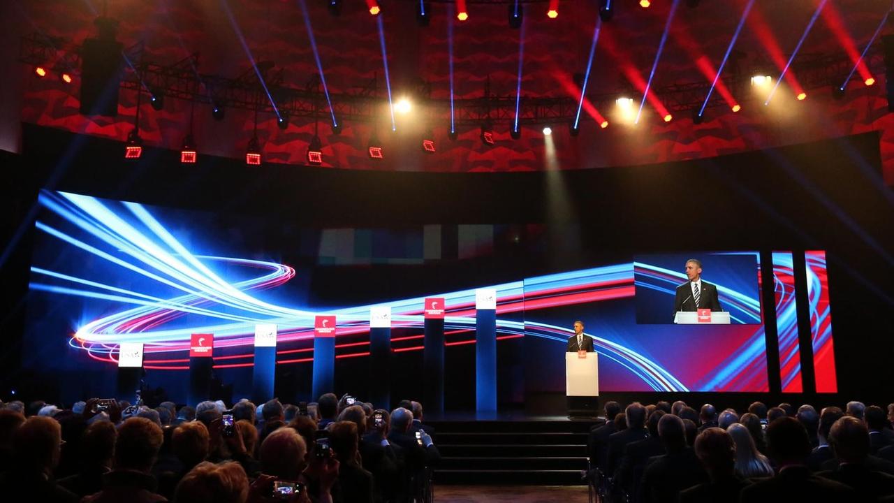 Sie sehen Barack Obama auf einer großen Bühne am Rednerpult, dahinter bunte Licht-Kurven als Beleuchtung. Sie sehen Barack Obama auf einer großen Bühne am Rednerpult, dahinter bunte Licht-Kurven als Beleuchtung.